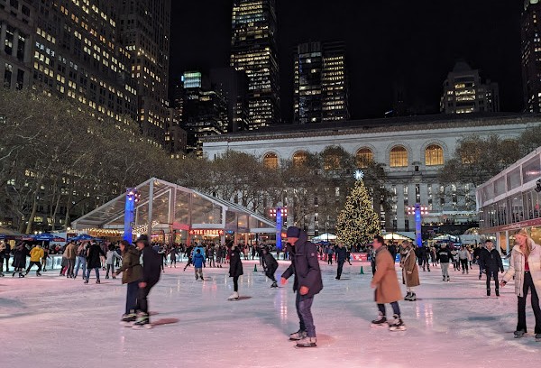 Persone che pattinano di notte sul giacchio sulla pista illuminata del Winter Village. Grattacieli di New York sullo sfondo e alberi di Natale